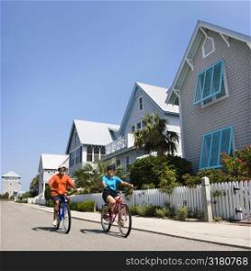 Middle aged African American couple bicycling down street next to homes and fence.