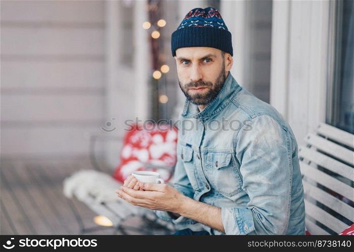 Middle aged bearded male with blue eyes, thick beard and mustache, wears stylish hat and denim jacket, holds cup of hot tea or coffee, has thoughtful expression, thinks about something important