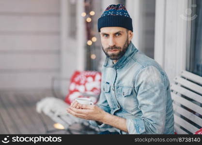 Middle aged bearded male with blue eyes, thick beard and mustache, wears stylish hat and denim jacket, holds cup of hot tea or coffee, has thoughtful expression, thinks about something important