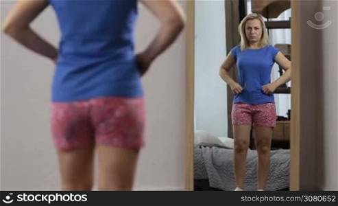Middle-aged blonde female looking at her reflection in the mirror, upset and dissatisfied with her body image at home. Displeased woman pinching her stomach fat while looking at the mirror.