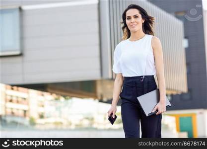 Middle-aged businesswoman working with her smart phone and laptop outdoors. Woman walking near business building with very careful hair.. Middle-aged businesswoman working with her smart phone and laptop outdoors.