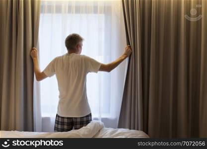 Middle aged Caucasian man opening curtains looking through window in a hotel room beginning of a new day