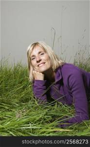 Middle-aged Caucasian woman lying in grass resting head on hand smiling.