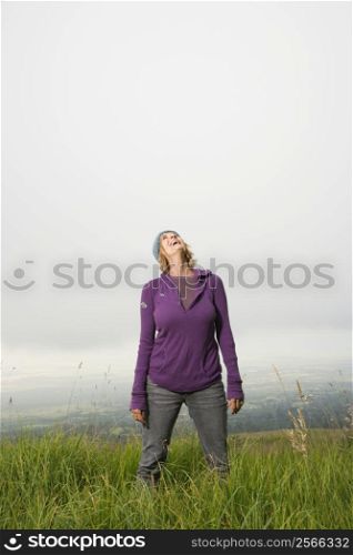 Middle-aged Caucasian woman standing alone in field with head back laughing.