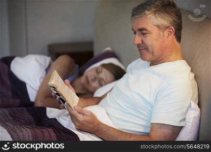 Middle Aged Couple In Bed Together With Man Reading Book