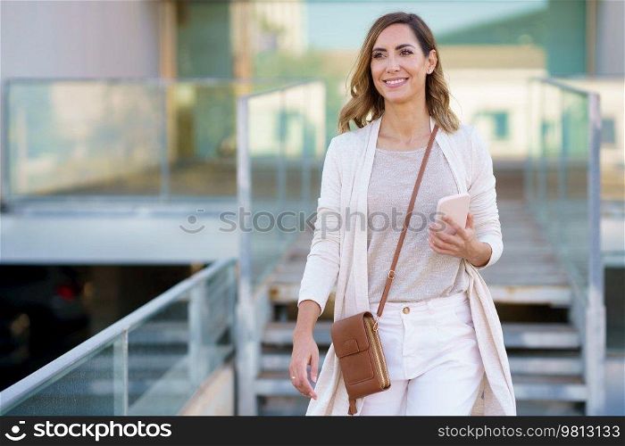 Middle aged female standing near an office building carrying a handbag and a smartphone. Caucasian woman in urban background.. Caucasian middle aged female standing near an office building carrying a handbag and a smartphone.