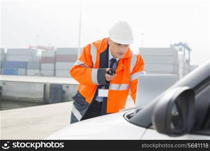 Middle-aged man talking on walkie-talkie while using laptop in shipping yard