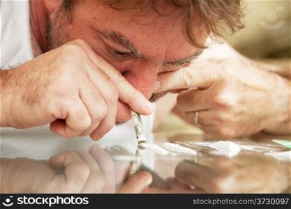 Middle-aged man using a rolled up dollar bill to snort cocaine off a glass coffee table. **Dramatization - no illegal drugs were used in the creation of this photo**