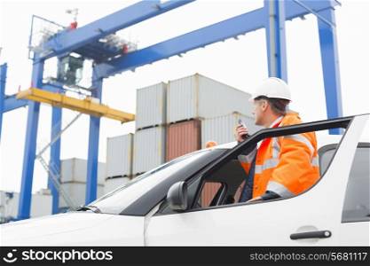 Middle-aged man using walkie-talkie while standing at car door in shipping yard