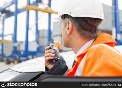 Middle-aged man using walkie-talkie while standing beside car in shipping yard