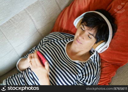 Middle-aged woman consulting her smartphone while listening to music with headphones lying on the couch.. Woman consulting her smartphone while listening to music with headphones lying on the sofa.
