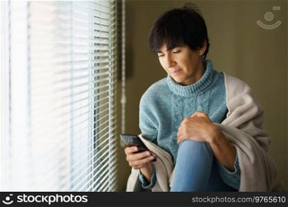 Middle-aged woman consulting something on her smartphone near the window at home. Female in her 50s. Middle-aged woman consulting something on her smartphone near the window at home.