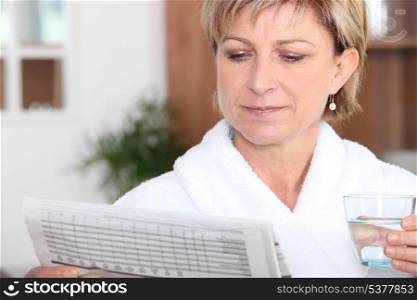 Middle-aged woman drinking glass of water and reading newspaper