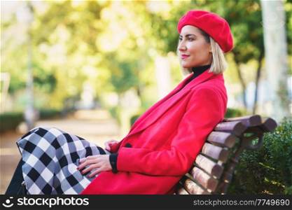 Middle-aged woman sitting on a bench in an urban park wearing red winter clothing. Female wearing coat, skirt and beret outdoors.. Middle-aged woman sitting on a bench in an urban park wearing red winter clothing.