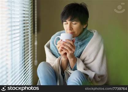 Middle-aged woman smelling coffee from her cup near a window. Female in her 50s. Middle-aged woman smelling coffee from her cup near a window.