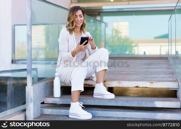 Middle aged woman taken a coffee break, sitting on steps near her office building with an eco-friendly ecological metal water bottle. Businesswoman using smartphone.. Middle aged woman using smartphone sitting on steps near her office. Famela taken a coffee break.