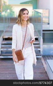 Middle-aged woman walking near an office building carrying a handbag and a smartphone. Caucasian female in urban background.. Middle-aged woman walking near an office building carrying a handbag and a smartphone.