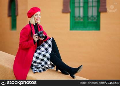 Middle-aged woman wearing red winter clothes, taking pictures with an SLR camera sitting on a city wall. Female wearing coat, skirt and beret outdoors.. Woman wearing red winter clothes, taking pictures with an SLR camera sitting on a city wall.