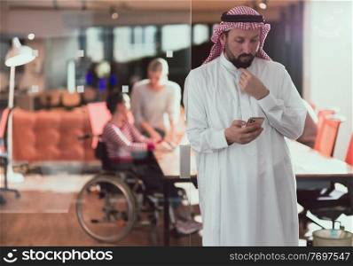 Middle eastern Arab businessman in the office meeting room speaking on the smartphone. Disabled businesswoman in wheelchair working together with the team in the background. High quality photo. Follow focus. . Middle eastern Arab businessman in the office speaking on smartphone in front of his team 