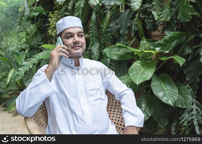 Middle eastern Emirati man in traditional clothing called kandura sitting on chair in coffee shop and talking with his mobile.Smart communication technology concept.