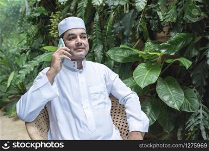 Middle eastern Emirati man in traditional clothing called kandura sitting on chair in coffee shop and talking with his mobile.Smart communication technology concept.