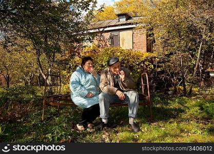 Middleaged man and woman sit on old rusty bed in autumnal garden, eat apple and smoke