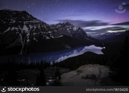 Midnight at Peyto Lake where a hiker lights the shore below and faint aurora greens and purples shine to the north.
