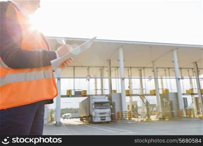 Midsection of female worker writing on clipboard while looking at truck entering in shipping yard