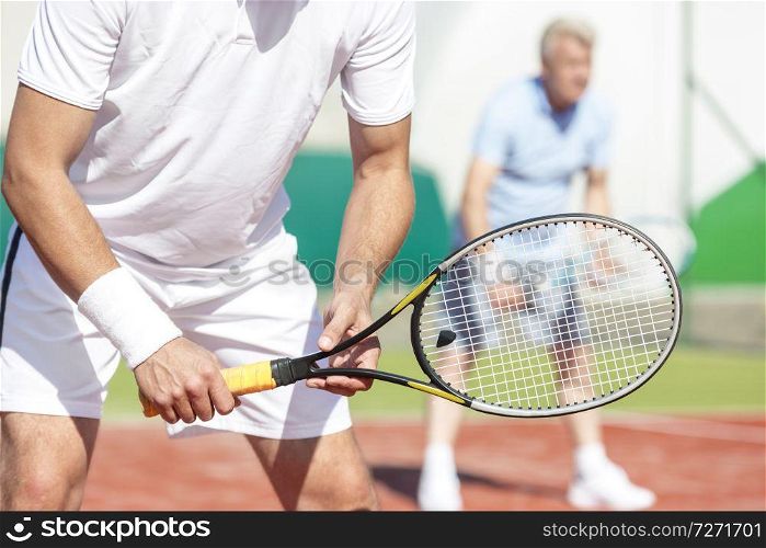 Midsection of man standing with tennis racket against friend playing doubles match on court