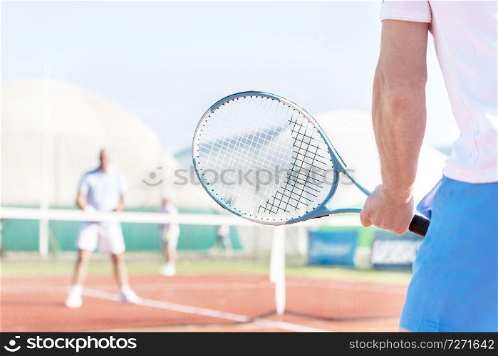 Midsection of mature man holding racket while playing with friends on tennis court