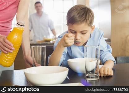 Midsection of woman with juice bottle standing by son having breakfast with man in background