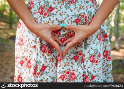 Midsection of young woman in white summer dress with flower print holding her hands together in triangular shape with fingers and thumbs touching in the lower belly genital region