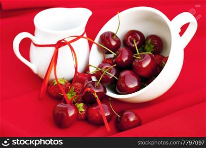 milk jug with a ribbon cherries and strawberries in a white cup on a red background