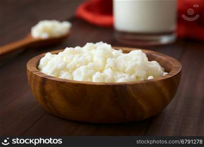 Milk kefir grains in wooden bowl with a glass of kefir in the back, photographed with natural light (Selective Focus, Focus one third into the kefir grains)