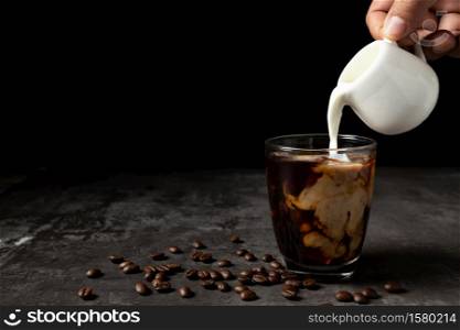 Milk pouring into iced black coffee on table and black background.