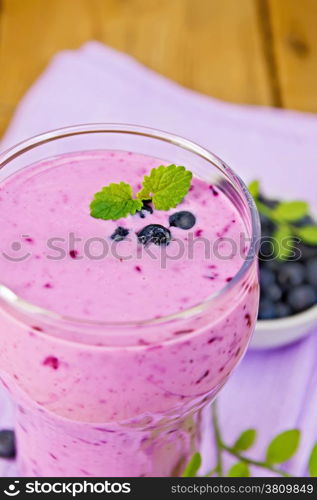 Milkshake with blueberries in a glass on a purple napkin, saucer with blueberries on a wooden boards background