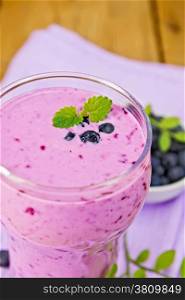 Milkshake with blueberries in a glass on a purple napkin, saucer with blueberries on a wooden boards background