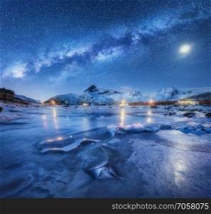 Milky Way above frozen sea coast, snow covered mountains and starry sky with moon in winter at night in Norway. Landscape with galaxy, ice, snowy rocks, buildings, illumination, milky way. Space