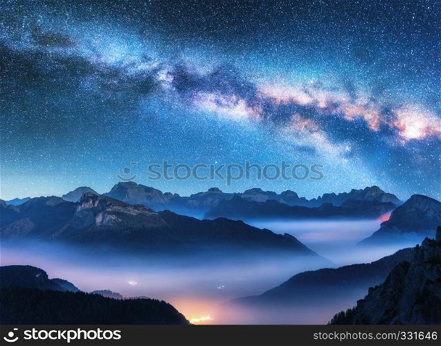 Milky Way above mountains in fog at night in summer. Landscape with alpine mountain valley, low clouds, purple starry sky with milky way, city illumination. Aerial. Passo Giau, Dolomites, Italy. Space