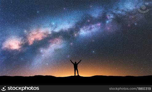 Milky Way and happy man at night. Silhouette of alone guy with raised up arms, sky with bright stars, orange light in summer. Galaxy. Space background. Landscape with milky way. Travel and nature