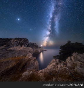 Milky Way and lighthouse on the mountain peak at starry night in summer. Beautiful landscape with cliffs, rocky sea coast, sky with bright milky way and stars. Space and nature. Cape Lefkada, Greece. Milky Way and lighthouse on the mountain peak at starry night