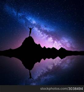 Milky Way and silhouette of a standing woman on mountain. Milky Way. Night starry sky and silhouette of a standing woman with raised up arms on the mountain peak near the lake with reflection in water. Landscape with Milky Way and woman. Galaxy, Universe