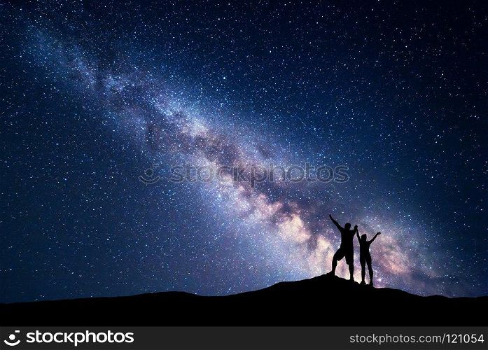Milky Way. Night sky with stars and silhouette of standing happy man and woman with raised  up arms on the hill. Blue milky way with people on the mountain. Background with universe. Night landscape