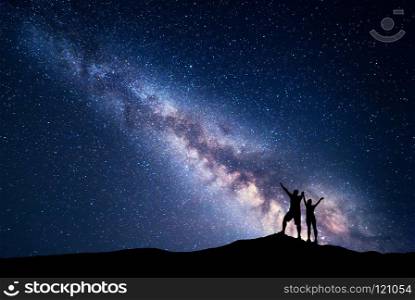 Milky Way. Night sky with stars and silhouette of standing happy man and woman with raised  up arms on the hill. Blue milky way with people on the mountain. Background with universe. Night landscape