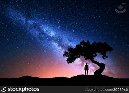 Milky Way. Night sky with stars, old tree and silhouette of a standing alone man on the mountain. Milky way with red light and man on the hill. Galaxy. Silhouette of a man under the tree. Universe