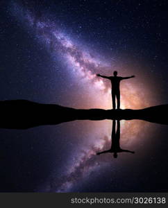 Milky Way. Night starry and silhouette of a standing alone man with with raised up arms on the mountain near the lake with sky reflection in water. Milky way and man on the hill. Galaxy. Universe