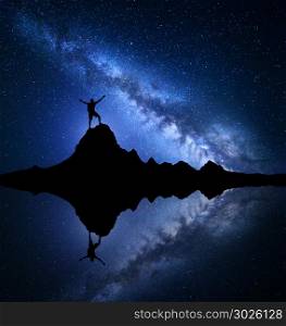 Milky Way. Night starry sky and silhouette of a standing happy man with raised up arms on the mountain peak near the lake with reflection in water. Blue milky way and man on the rock. Galaxy. Universe