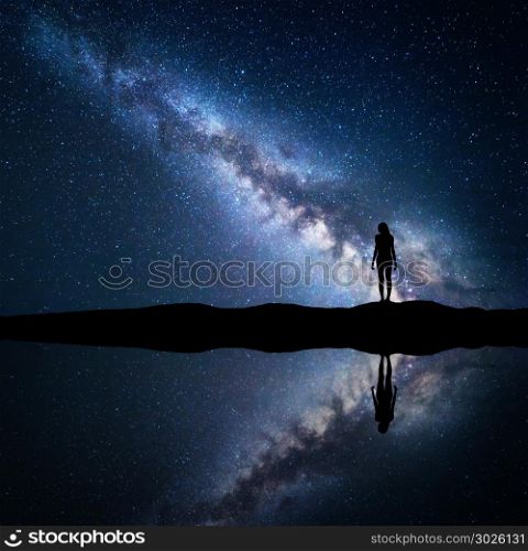 Milky Way. Night starry sky and silhouette of a standing woman on the mountain near the lake with sky reflection in water. Landscape with blue Milky Way and woman. Galaxy, Universe. Space background.