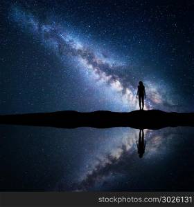 Milky Way. Night starry sky and silhouette of a standing woman on the mountain near the lake with sky reflection in water. Landscape with blue Milky Way and woman. Galaxy, Universe. Space background.