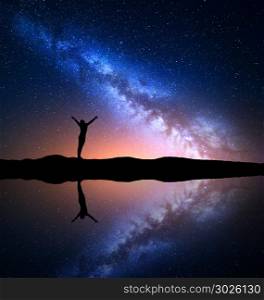 Milky Way. Night starry sky and silhouette of a standing woman with raised up arms on the hill near the lake with reflection in water. Colorful landscape with Milky Way and woman. Universe. Space